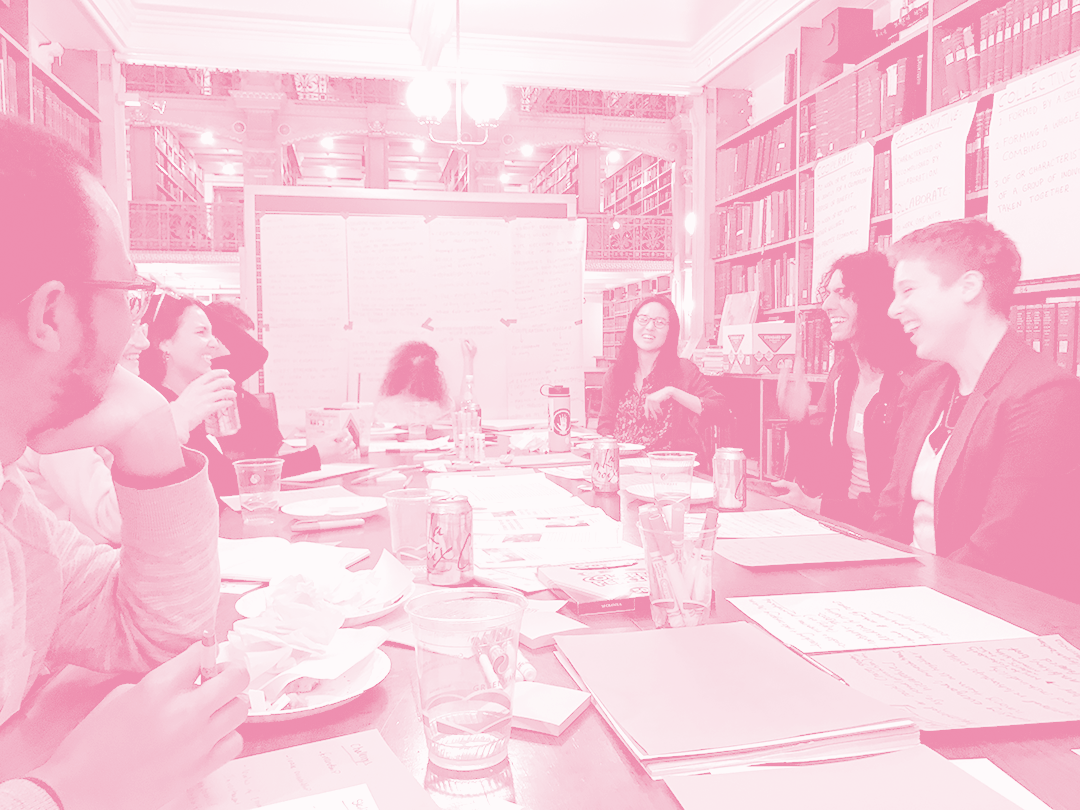 A two-tone green and white photograph of a group of people sitting around a large table that is covered in pieces of paper with handwritten notes, cups, a disposable plate and a can of soda, look across the table from one another laughing. Behind two people who are sitting on the right side is a big shelf of books that covers the entire wall. At the end of the table is a chalkboard covered with large sheets of newsprint with writing on them and a person writing notes at the bottom of the board.